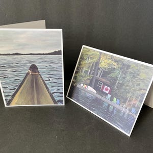 Könnte beinhalten: Zwei Grußkarten mit malerischen Seenszenen. Die erste Karte zeigt ein Kanu auf einem See mit einem Wald im Hintergrund. Die zweite Karte zeigt eine Hütte an einem See mit Bäumen und bunten Stühlen.