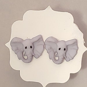 May include: A pair of light purple elephant-shaped stud earrings with black eyes. The earrings are displayed on a white, scalloped-edge card against a neutral background. The elephant earrings are a cute and whimsical accessory.