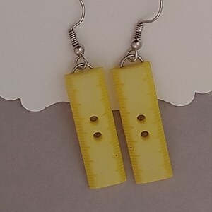 Puede incluir: Un par de pendientes amarillos con forma de botón y ganchos plateados.