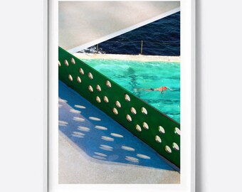 May include: A green metal fence with circular cutouts casts shadows on a concrete surface. A swimming pool with turquoise water is visible through the fence, with a person swimming in the distance.