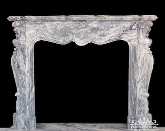 May include: A white marble fireplace mantel with intricate carvings and a scalloped top. The mantel is freestanding and has a rectangular opening for a fireplace.