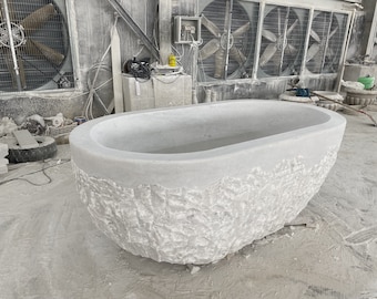 May include: A large, white, oval-shaped bathtub made of stone with a rough, textured exterior. The bathtub is sitting on a concrete floor in a workshop setting.