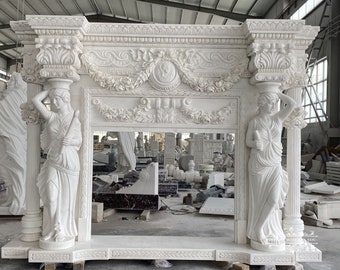 May include: A white marble fireplace mantel with intricate carvings and two statues of women on either side. The mantel is in a workshop setting with other unfinished marble pieces in the background.