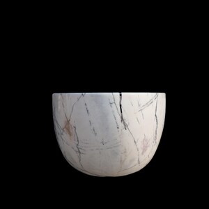 May include: A white marble bowl with gray and brown veining. The bowl has a rounded shape and a smooth surface. The background is solid black. This decorative bowl is suitable for home decor.