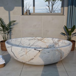 May include: A large, oval-shaped marble bathtub with a white and gray veined pattern. The tub is set in a bathroom with potted plants and a window. The tub's dimensions are approximately 72 inches long and 36 inches wide.