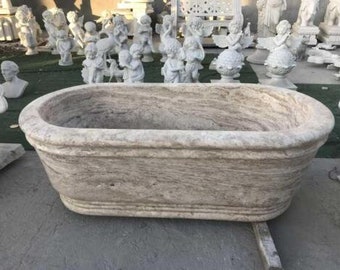 May include: A large, oval-shaped, stone bathtub with a light brown and white marbled finish. The bathtub is sitting on a concrete surface.