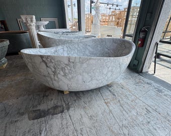 May include: A large, oval-shaped marble bathtub with gray veining. The tub is set on a weathered wooden floor, with a view of the outdoors through a large window. Other marble fixtures are visible in the background.