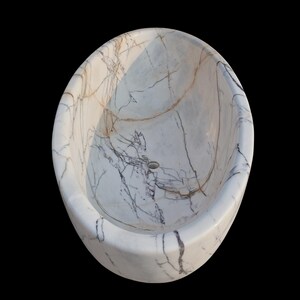 May include: Oval-shaped white marble bathtub with gray and brown veining. The freestanding tub has a smooth, polished surface and a drain hole at the bottom. The design is a modern take on a classic bathroom fixture.
