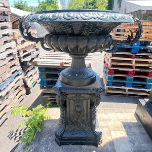 May include: Black cast iron planter with a decorative base and a large, ornate bowl. The planter is sitting on a wooden pallet.