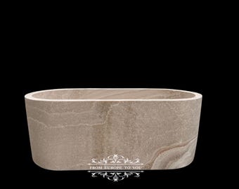 May include: A beige, oval-shaped stone bathtub with a smooth, polished finish. The tub has a rectangular shape with rounded edges and a thick rim. The stone has natural veining and a neutral color palette, suitable for a modern bathroom.