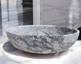 May include: Oval marble sink with grey and white veining. The vessel sink sits on a white marble countertop. The bathroom fixture has a smooth, polished finish and a natural stone appearance. The sink is a modern design.