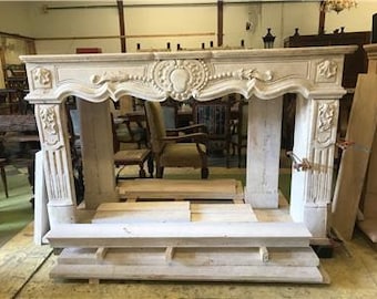 May include: A white marble fireplace mantel with ornate carvings and a decorative design. The mantel is in a workshop setting and is resting on wooden supports.