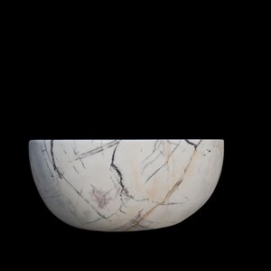 May include: A white marble bowl with dark gray and light brown veining. The bowl has a rounded, semi-circular shape and sits against a black background. The bowl's surface has a polished finish.