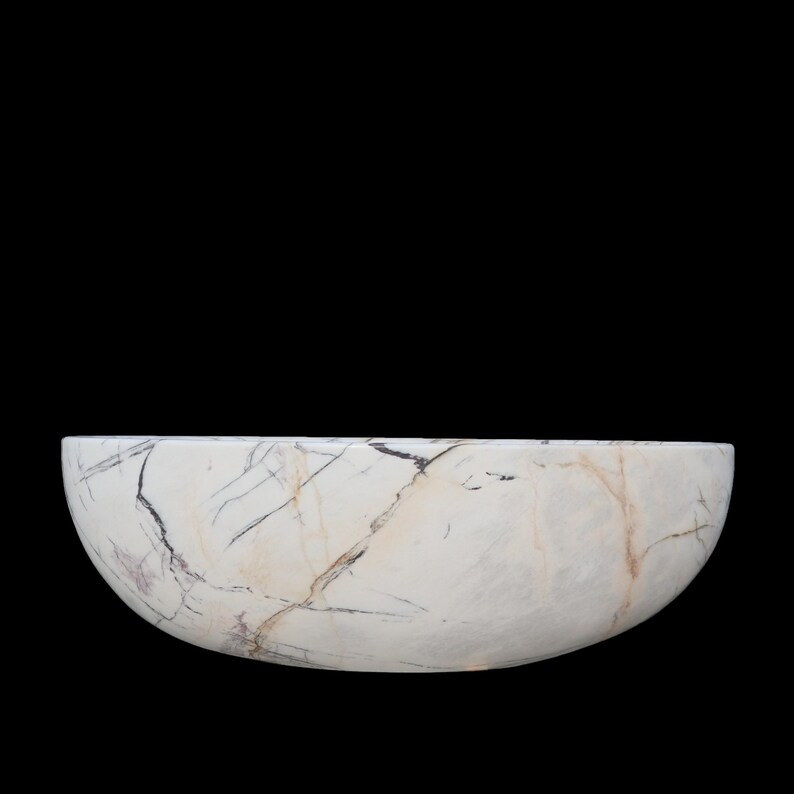 May include: A white marble bowl with a semi-circular shape. The bowl has dark gray and brown veining throughout the surface. The background is solid black.