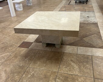 May include: A square, light beige, stone coffee table with a simple, modern design. The table has a thick, flat top and a square base with rounded edges. The table is sitting on a tiled floor.