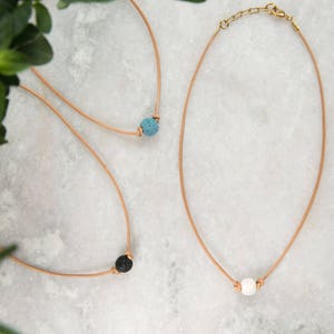 Puede incluir: Tres collares de cuero con cuentas de roca volcánica. Los collares están sobre una superficie de mármol blanco. Las cuentas son azules, negras y blancas.