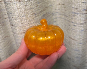 Puede incluir: Un pequeño objeto decorativo en forma de calabaza naranja con un acabado brillante. La calabaza tiene un tallo curvo y se sostiene en una mano. Es probable que sea una decoración de temporada, perfecta para el otoño o Halloween.