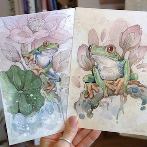Könnte beinhalten: Zwei Aquarellmalereien von Laubfröschen. Die Frösche sind grün mit roten Augen und sitzen auf Seerosenblättern und Lotusblüten. Der Hintergrund ist eine weiche Aquarellwäsche aus Rosa und Blau. Das Kunstwerk ist detailliert und skurril.
