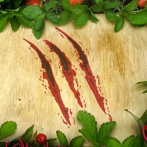 May include: Three red claw marks on a wooden background surrounded by green leaves and red strawberries.