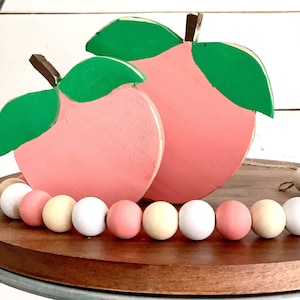 May include: Two wooden peach shaped ornaments with green leaves, sitting on a wooden tray with a string of pastel colored wooden beads.