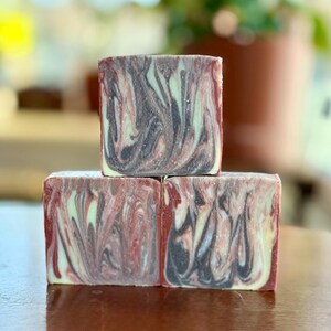May include: Three square bars of handmade soap stacked on top of each other. The soap has a marbled design with swirls of red, black, and pale green. The soap is sitting on a wooden surface.