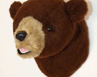 May include: A brown bear head wall mount with a light brown muzzle and pink tongue. The bear has dark eyes and a black nose. The fur is a soft, plush material. This decorative item is designed to be mounted on a wall.