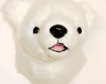 May include: A plush wall-mounted polar bear head featuring soft white faux fur, round ears, dark plastic eyes, a black nose, and a slightly open mouth revealing a pink tongue. The design captures the facial features of a polar bear in detail.