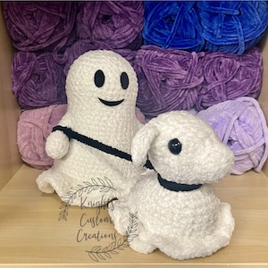 May include: A white crocheted ghost holding a black leash attached to a white crocheted lamb. The ghost has a black smiling face. The ghost and lamb are sitting on a wooden surface with purple and blue yarn in the background.