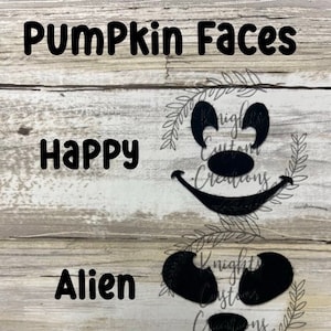 May include: Two black pumpkin face cutouts on a wood background. The top cutout is a happy face with a wide smile and large eyes. The bottom cutout is an alien face with large, round eyes and a small nose.