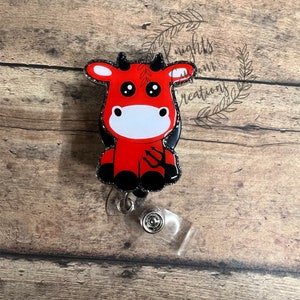 May include: Red glitter devil cow cartoon badge reel with black horns, eyes, and hooves. The reel is clear plastic with a silver metal clip.