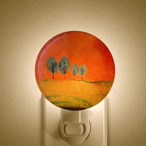 May include: A round night light with a landscape painting. The artwork features four blue-green trees on a yellow field under an orange sky. The night light is plugged into a white wall outlet.