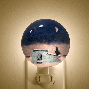 May include: A nightlight featuring a blue and white winter scene. The scene depicts a small house with a lit window, a pine tree, and a crescent moon in the sky.