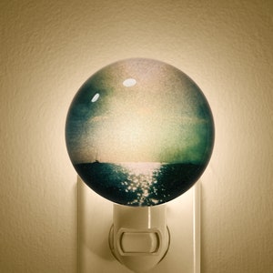 May include: A round, white night light with a picture of a sunset over the ocean. The night light is plugged into an outlet.