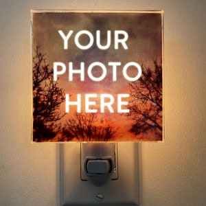 Peut inclure: Veilleuse carrée avec l'inscription blanche "YOUR PHOTO HERE", éclairée sur un fond de coucher de soleil avec des arbres en silhouette. La veilleuse se branche sur une prise murale standard.