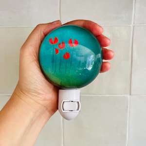 Peut inclure: Une veilleuse bleu turquoise avec un interrupteur blanc et un motif peint de fleurs rouges. La veilleuse a la forme d'une sphère.