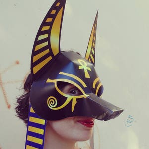 May include: A black and gold paper mask with an Ankh symbol and a stylized eye design. The mask has two large ears and is worn by a person with a blue and gold striped scarf.