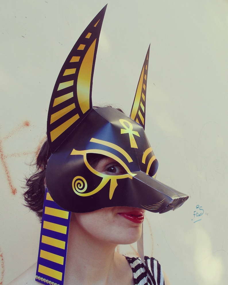 May include: A black and gold paper mask with an Ankh symbol and a stylized eye design. The mask has two large ears and is worn by a person with a blue and gold striped scarf.