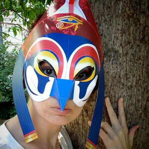 May include: A person wearing a colorful paper mask with a blue, red, and yellow design. The mask has a large eye on the forehead and a long, pointed beak. The mask is held in place by two blue straps that tie behind the head.