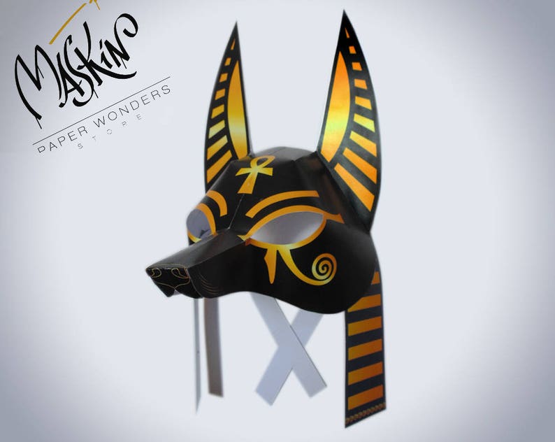 May include: A black and gold paper mask of an Anubis jackal head with a white eye cutout. The mask has a long, striped, gold and black strap.