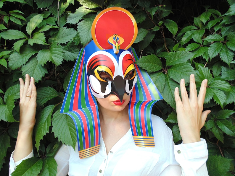 May include: A colorful paper mask with a red, blue, green, and yellow design. The mask has a bird-like beak and a crown on top. The mask is worn by a person with a white shirt.