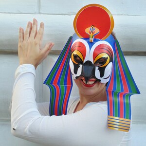 May include: A woman wearing a colorful paper mask with a red, white, and blue design. The mask has a large, round, red headpiece and a long, pointed beak. The woman is wearing a white shirt and has her hand raised.