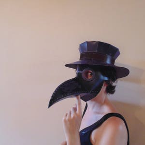 May include: A person wearing a black and purple plague doctor mask with a black hat and a black and white patterned dress.