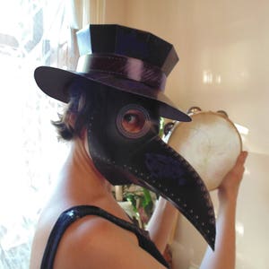 May include: A person wearing a black top hat and a black plague doctor mask is holding a tambourine. The mask has a long beak and a round eye hole. The person is wearing a black top hat with a black band around it.