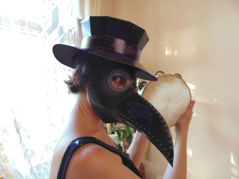 May include: A person wearing a black top hat and a black plague doctor mask is holding a tambourine. The mask has a long beak and a round eye hole. The person is wearing a black top hat with a black band around it.