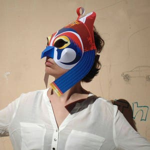 May include: A person wearing a colorful paper mask with a blue, red, and white design. The mask has a long, pointed beak and a crown on top. The person is wearing a white shirt.