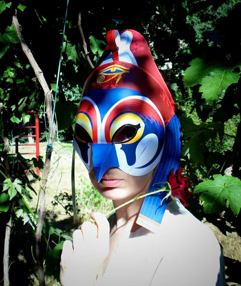 May include: A person wearing a blue, red, and yellow paper mask with a white eye design. The mask has a long, pointed nose and a white, blue, and red design on the side. The person is holding a red flower.