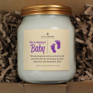 May include: A glass jar with a gold lid and a cream-colored label. The label has purple text that reads "We're Having A Baby" with two purple footprints. The text on the label also says "We are pregnant and so excited to tell you that John & I are having our first baby due 16th November 2022. www.sheenashona.com"