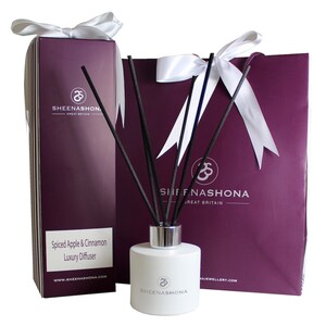 May include: A white bottle of Spiced Apple & Cinnamon Luxury Diffuser with black diffuser sticks and a white ribbon bow. The bottle is labeled with the brand name "SHEENASHONA" and the website address "www.SHEENASHONA.COM". The bottle is packaged in a purple gift box with a white ribbon bow and a matching purple gift bag with a white ribbon bow.