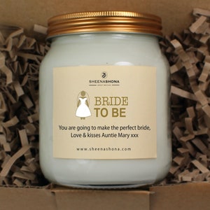 May include: A glass jar candle with a gold lid and a white label. The label has a gold graphic of a bride in a white wedding dress and the text "BRIDE TO BE". The text "You are going to make the perfect bride, Love & kisses Auntie Mary xxx" and "www.sheenashona.com" is also on the label.