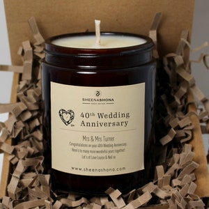 Puede incluir: Una vela en un tarro de vidrio marrón con una etiqueta blanca. La etiqueta tiene un diseño de corazón y el texto "40th Wedding Anniversary". El texto "Mrs & Mrs Turner" también está en la etiqueta. La vela está en una caja de regalo de papel marrón con papel triturado.